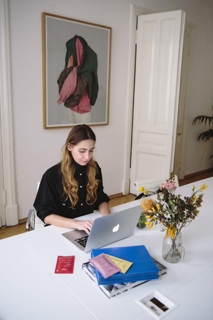 Young woman using a laptop at a desk with documents and flowers, in a bright, modern workspace.